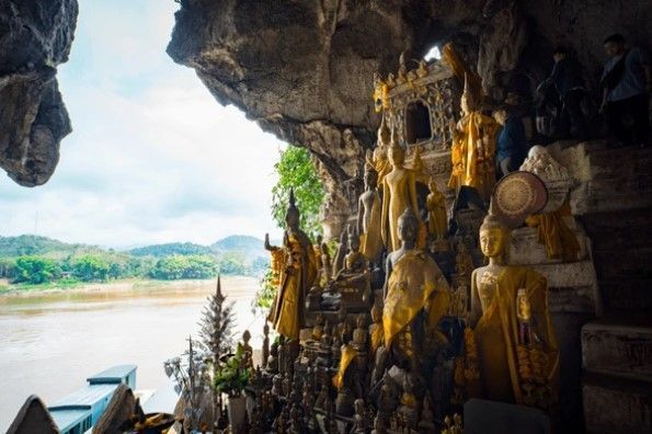 Un voyage en groupe WeRoad visitant une grotte remplie de statues de Bouddha dorées qui surplombe une large rivière et des collines verdoyantes.