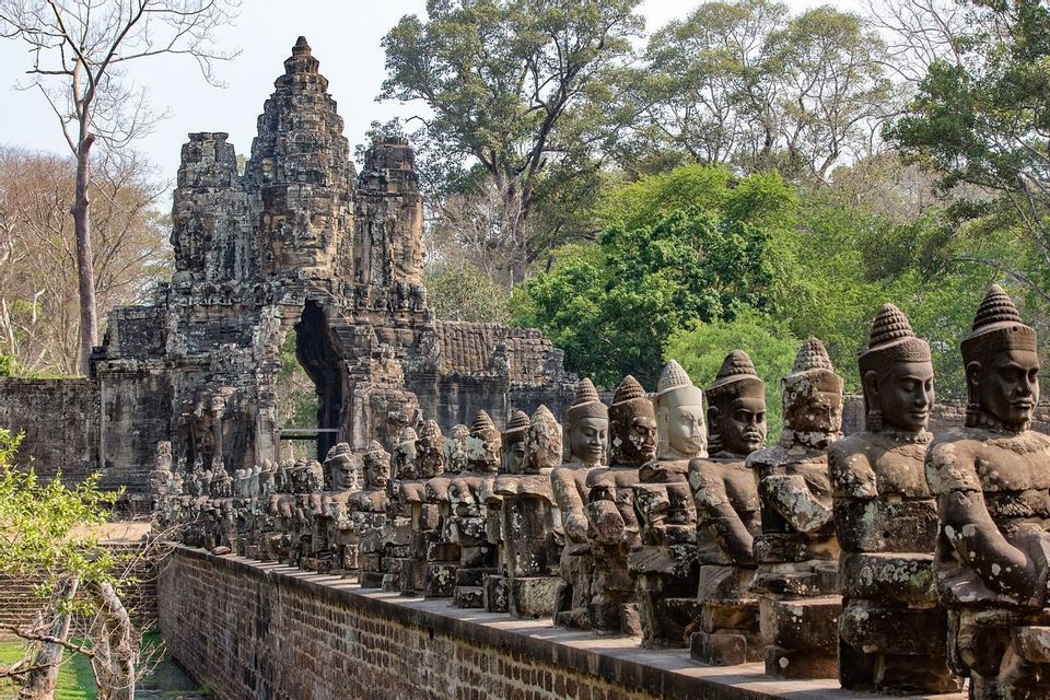 Une allée bordée de statues de pierre patinées conduit à une grande porte de temple sculptée, avec une forêt en toile de fond.