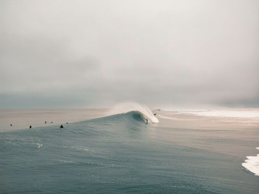 Un surfista cavalca un'enorme onda a tubo, mentre altri surfisti aspettano in acqua in una giornata nuvolosa.