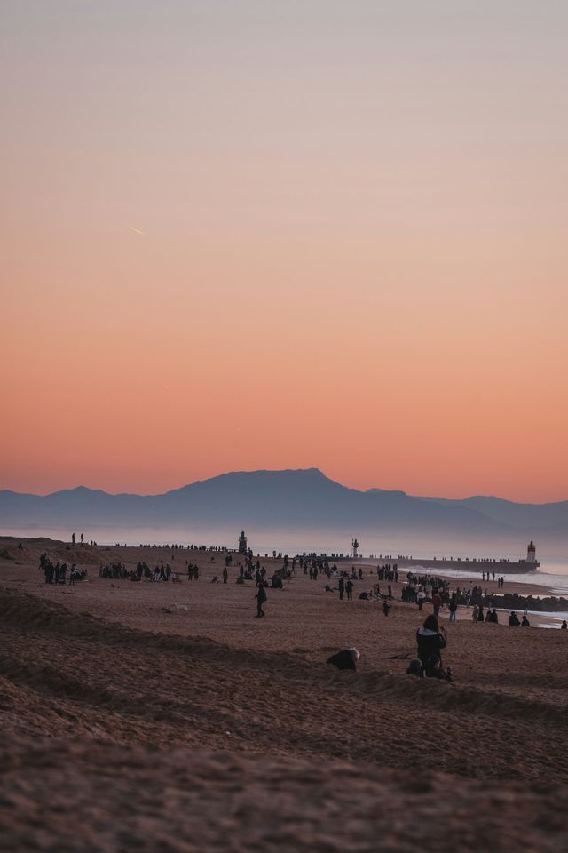 Una folla di persone si raduna su un'ampia spiaggia sabbiosa sotto un cielo arancione e velato al tramonto, con montagne e un molo in lontananza.