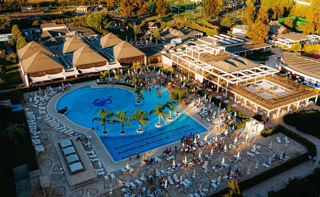 Vista aerea di un grande resort al tramonto, con piscina affollata, palme e edifici moderni.