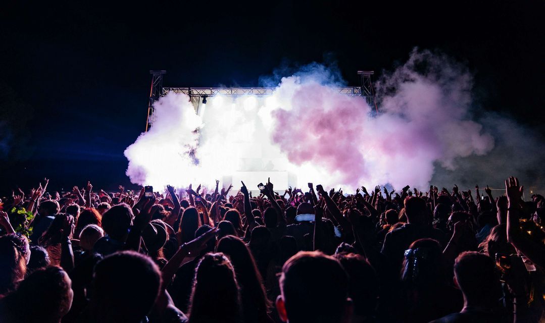 Una grande folla con le mani alzate di fronte a un palco illuminato a giorno, con fumo viola e bianco, durante un concerto notturno.