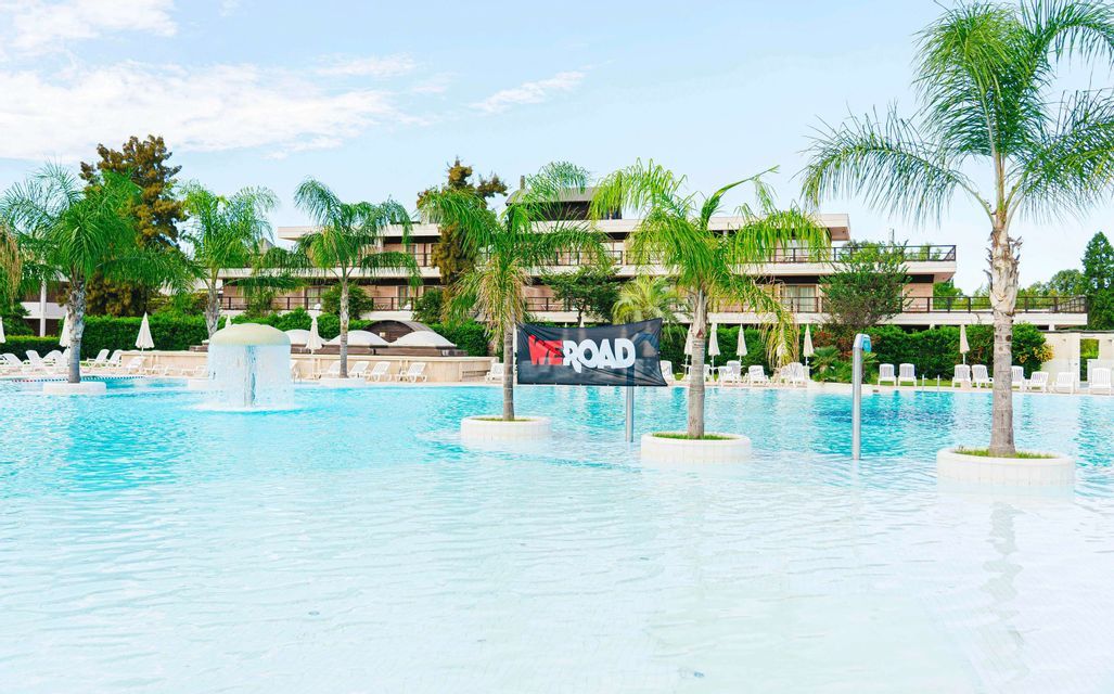 Un banner nero di WeRoad si trova in una grande piscina di un resort con una fontana, circondata da palme e sdraio.