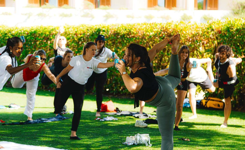 Un viaggio di gruppo WeRoad si dedica allo yoga della birra, bilanciandosi in posizioni su un prato verde mentre si beve dalle lattine.