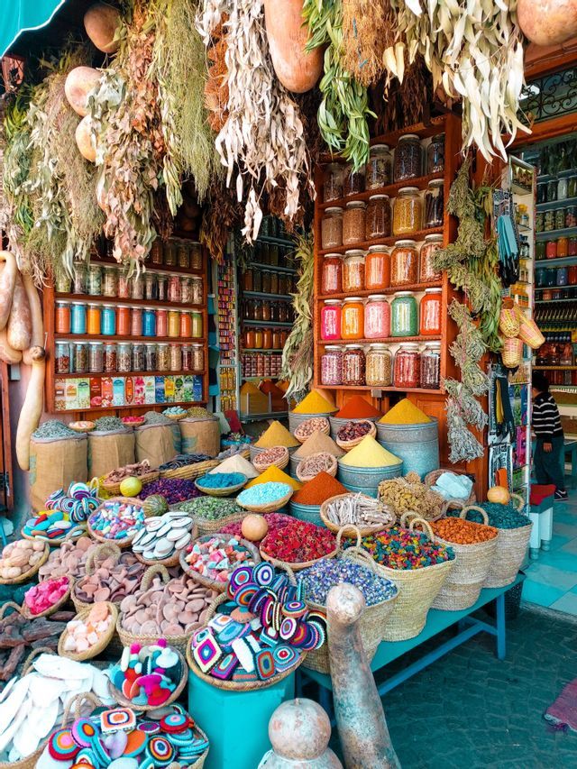 A market stall displays colorful spices in baskets and pyramids, with jars on shelves and dried herbs hanging from above.