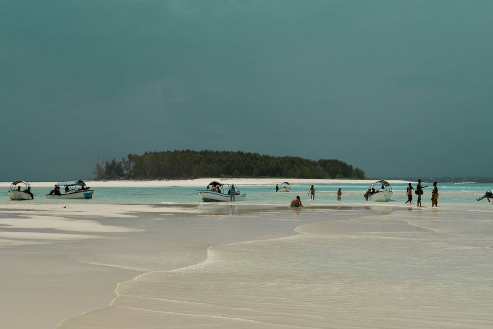 Un viaggio di gruppo WeRoad che guada in acque basse e turchesi con piccole barche su una spiaggia di sabbia bianca sotto un cielo coperto.