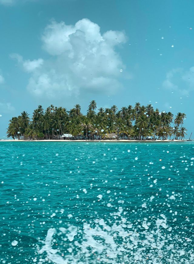 Un'isola tropicale coperta di palme vista dal mare turchese, con l'acqua che schizza in primo piano.