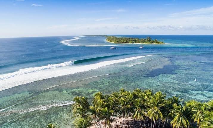 Veduta aerea di una lunga onda oceanica che si infrange lungo una barriera corallina turchese vicino a un'isola tropicale con palme e barche.