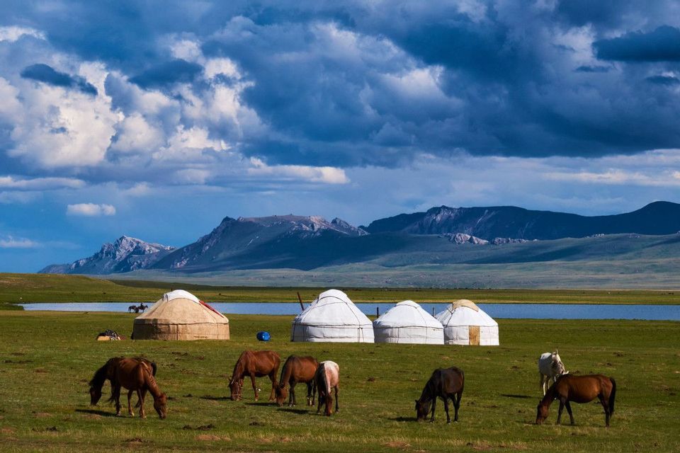 Pferde grasen auf einer grünen Weide, übersät mit Jurten, mit einem See und fernen Bergen unter bewölktem Himmel.