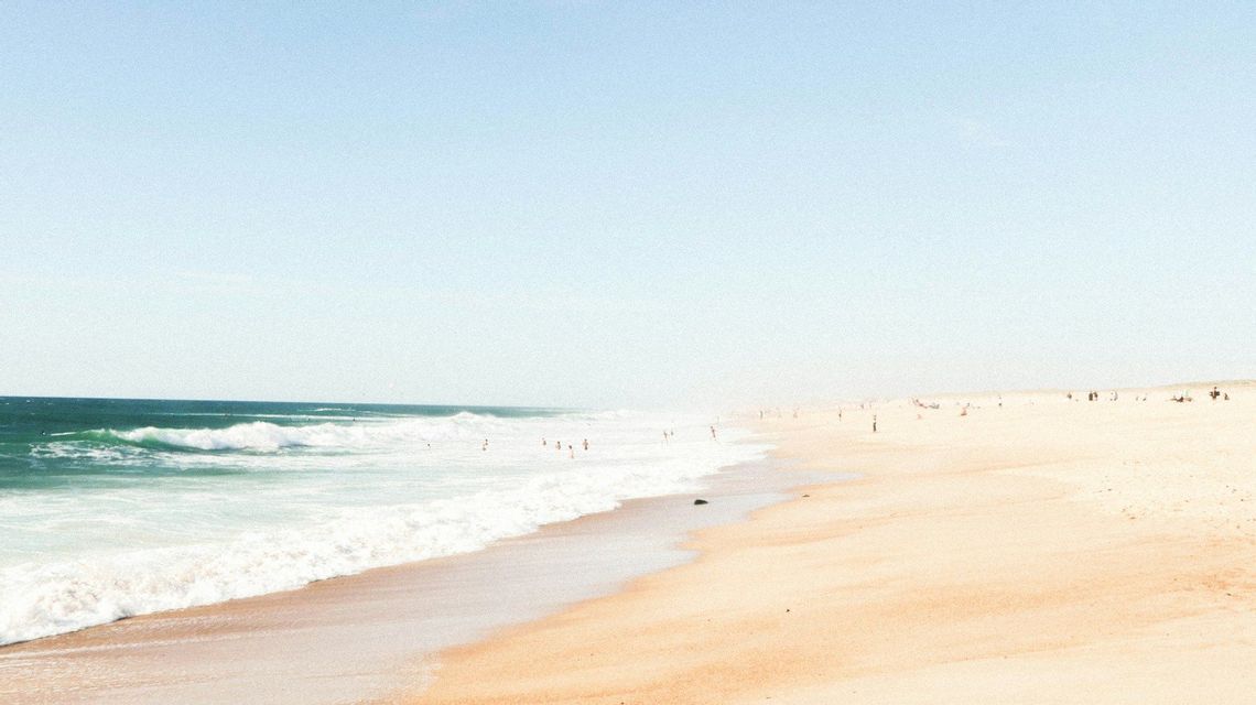Le onde dell'oceano lambiscono una vasta spiaggia sabbiosa, dove le persone nuotano e si rilassano sotto un cielo azzurro pallido.