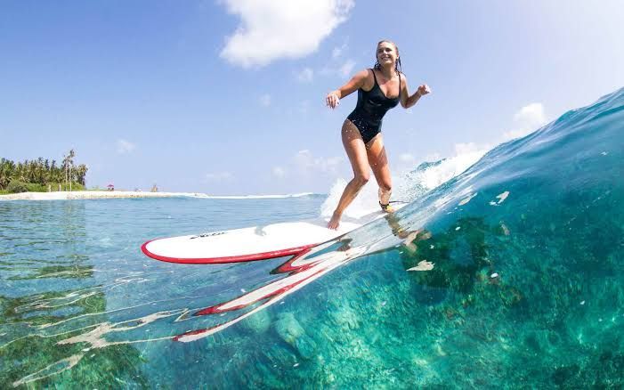 Una donna in costume nero fa surf su un'onda turchese trasparente, fotografata dal livello dell'acqua con una spiaggia tropicale sullo sfondo.