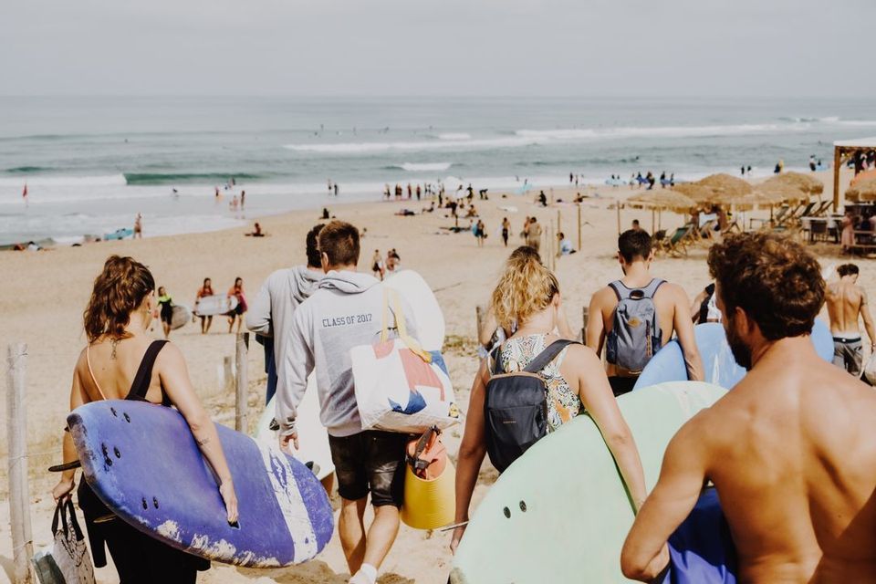 Un gruppo WeRoad, ripreso di spalle, cammina lungo un sentiero che porta a una spiaggia affollata con tavole da surf.