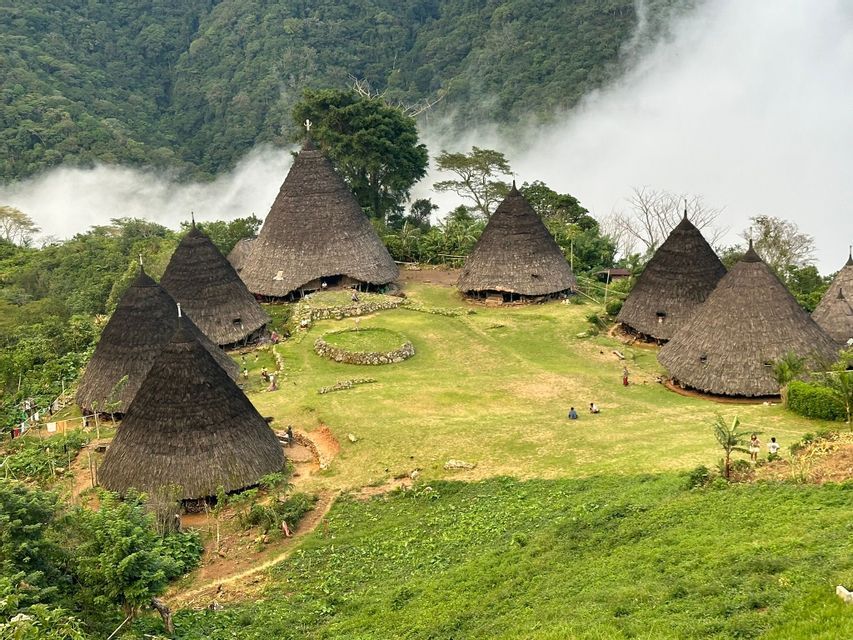 Un village traditionnel avec des huttes coniques aux toits de chaume sur une colline verdoyante, entouré de montagnes boisées et de nuages bas.