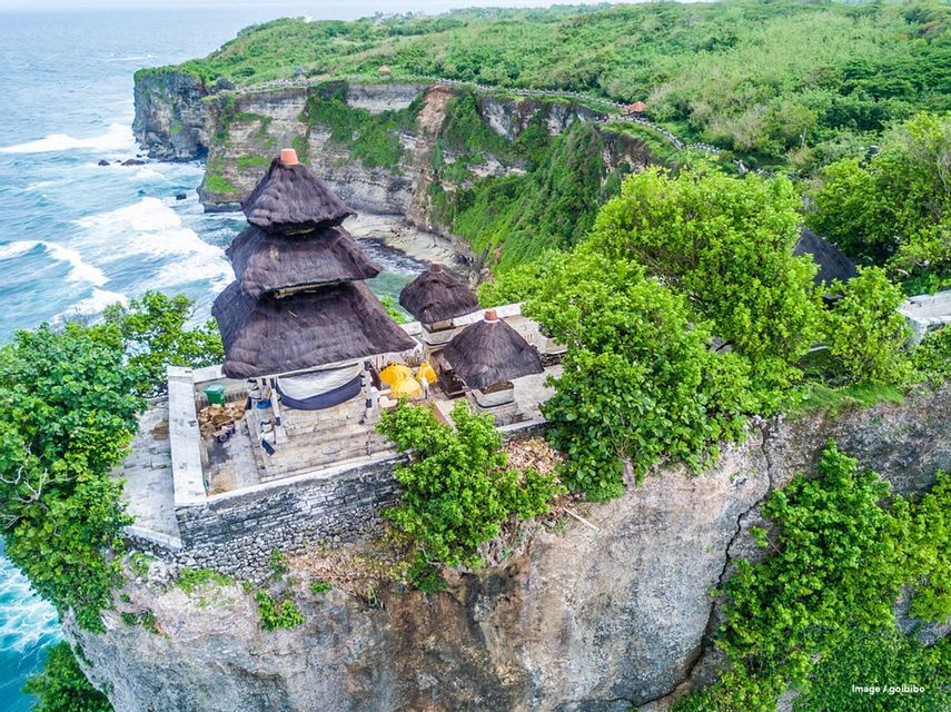 Une vue aérienne d'un temple traditionnel aux toits de chaume, perché sur une falaise verdoyante dominant l'océan.