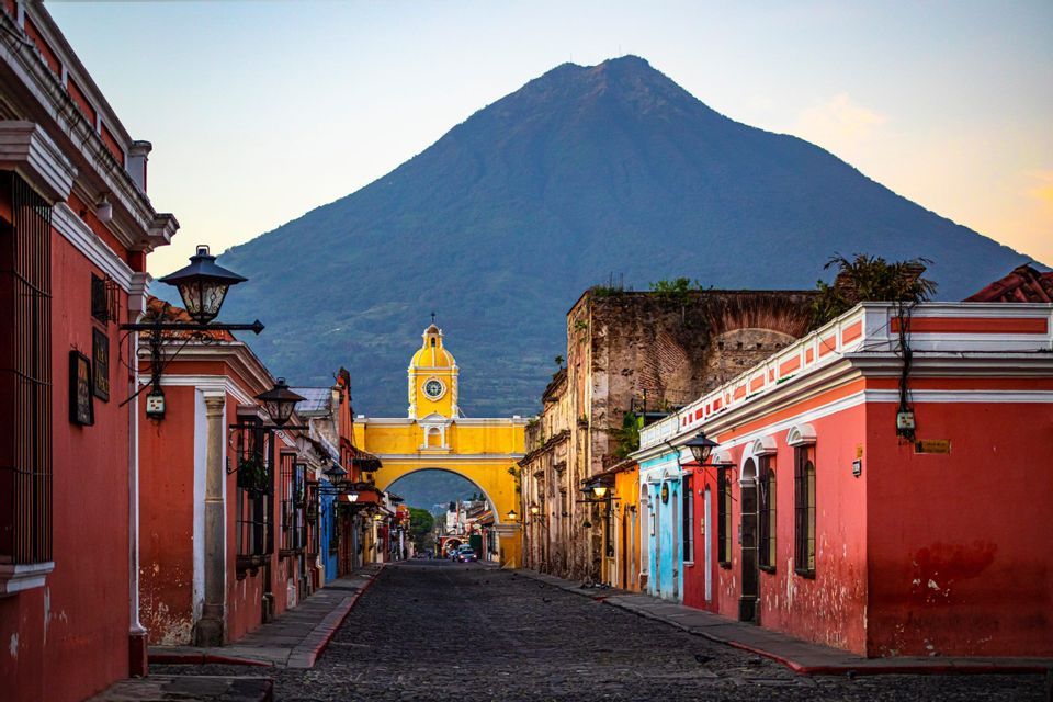Un arco giallo con una torre dell'orologio si erge alla fine di una strada acciottolata, con un grande vulcano che si staglia sullo sfondo.