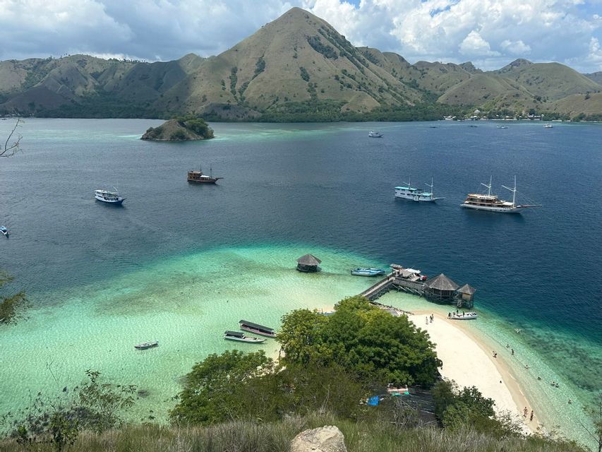 Vue plongeante sur des bateaux dans une baie tropicale avec une eau turquoise, une plage de sable blanc, un quai en bois et des collines verdoyantes.