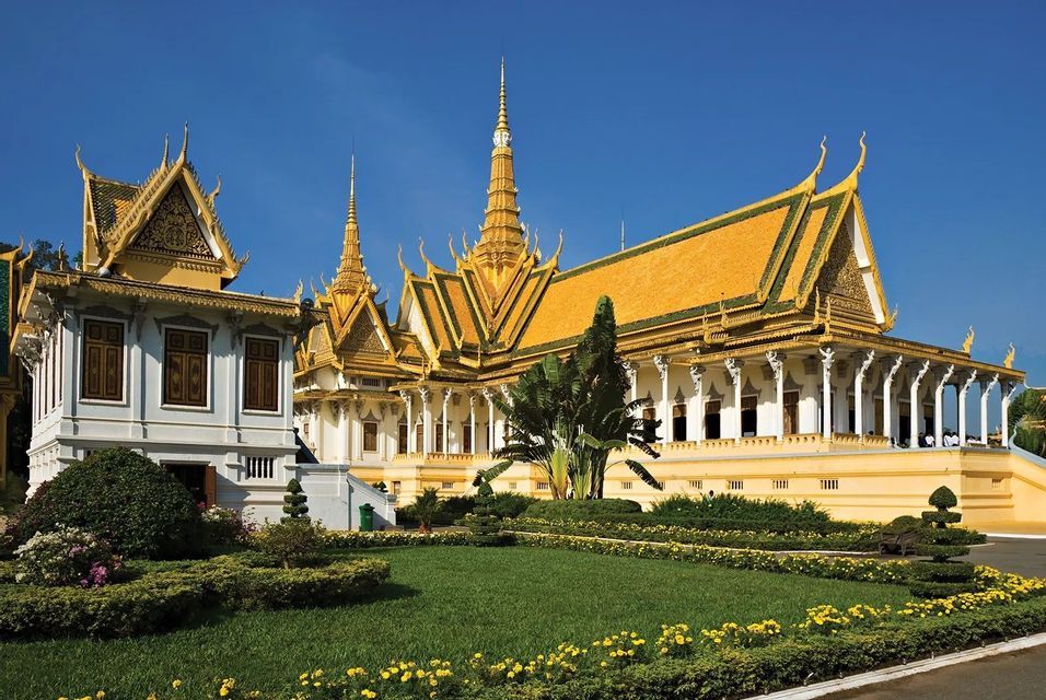 An ornate temple with golden roofs and spires sits behind a manicured green lawn with yellow flowers under a clear blue sky.
