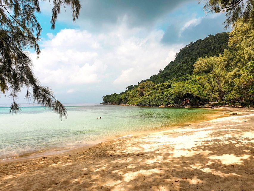 Ein Sandstrand mit Baumschatten führt zu ruhigem, klarem Wasser, wo zwei Personen neben einem bewaldeten Hügel schwimmen.