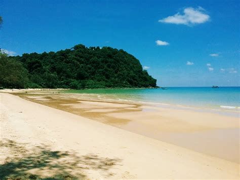 Ein breiter Sandstrand trifft auf ruhiges, türkisfarbenes Wasser neben einem üppigen, bewaldeten Hügel unter einem klaren blauen Himmel.