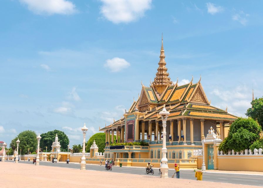 An ornate yellow palace with a multi-tiered golden roof stands beside a paved square under a bright blue sky.