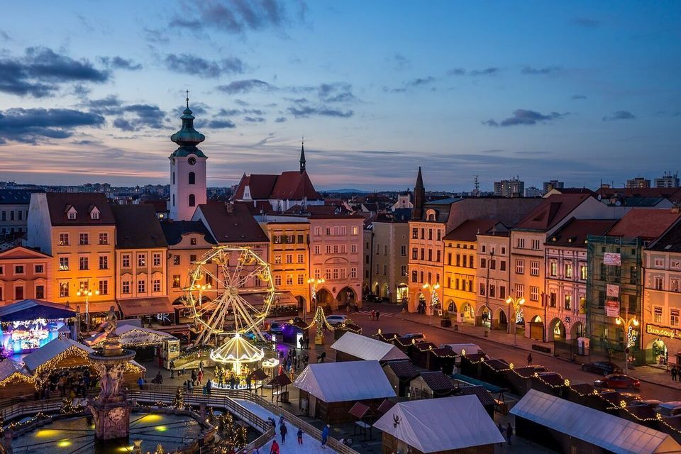 Luftaufnahme eines Weihnachtsmarkts auf einem Platz in der Abenddämmerung mit einem beleuchteten Riesenrad, Ständen und Menschen auf einer Eislaufbahn.