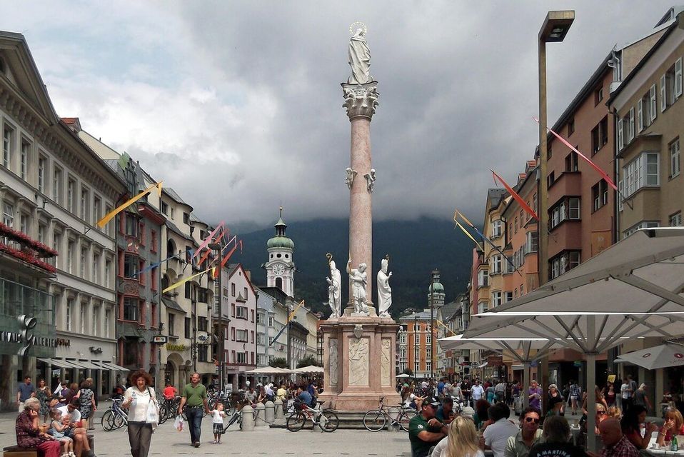 Auf einem belebten, von Menschen überfüllten europäischen Platz steht ein hohes, säulenförmiges Denkmal, unter einem bewölkten Himmel sind Berge zu sehen.