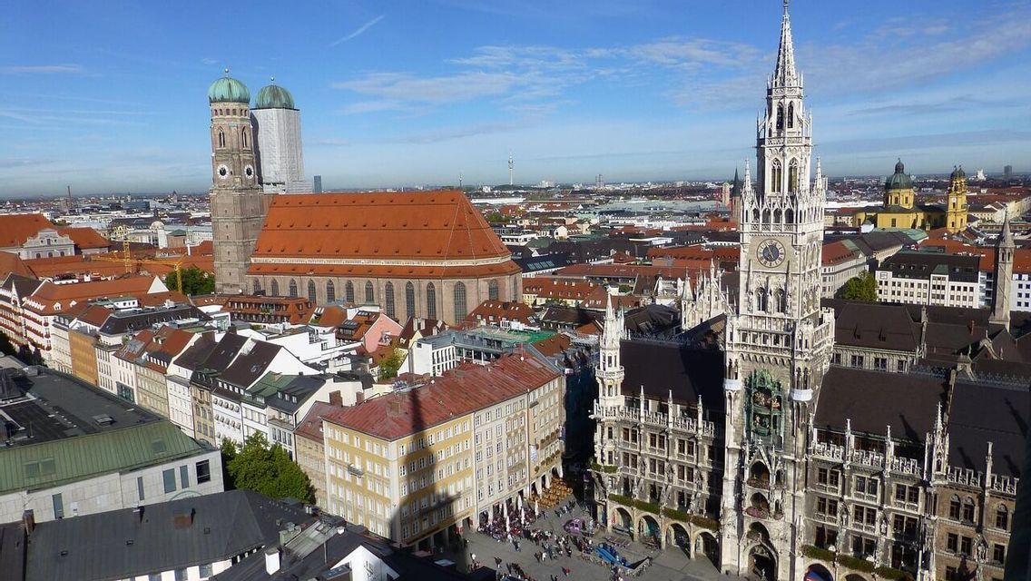 Eine Luftaufnahme einer europäischen Stadtlandschaft mit einem großen gotischen Uhrenturm und einer Kathedrale mit zwei Kuppeln unter blauem Himmel.