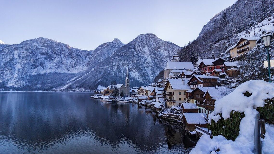 Ein schneebedecktes Dorf mit einer Kirche, eingebettet an einem ruhigen Seeufer am Fuße hoher Berge unter einem klaren Himmel.