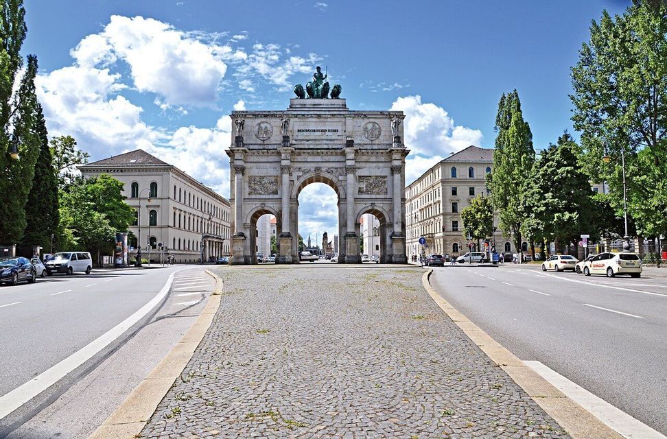 Eine breite, gepflasterte Straße führt zu einem großen, kunstvollen Triumphbogen, der von Gebäuden und Bäumen unter einem bewölkten blauen Himmel flankiert wird.
