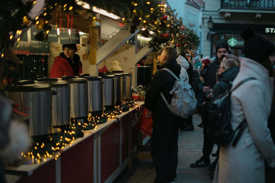 Eine Menschenmenge steht an einem mit Lichtern und Girlanden geschmückten Weihnachtsmarktstand im Freien Schlange, um heiße Getränke aus Stahlspendern zu bekommen.