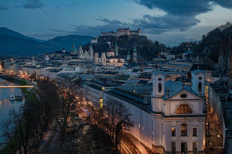 Eine bei Sonnenuntergang beleuchtete Stadtlandschaft mit einer Festung auf einem Hügel, historischen Gebäuden und Bergen in der Ferne unter einem bewölkten Himmel.
