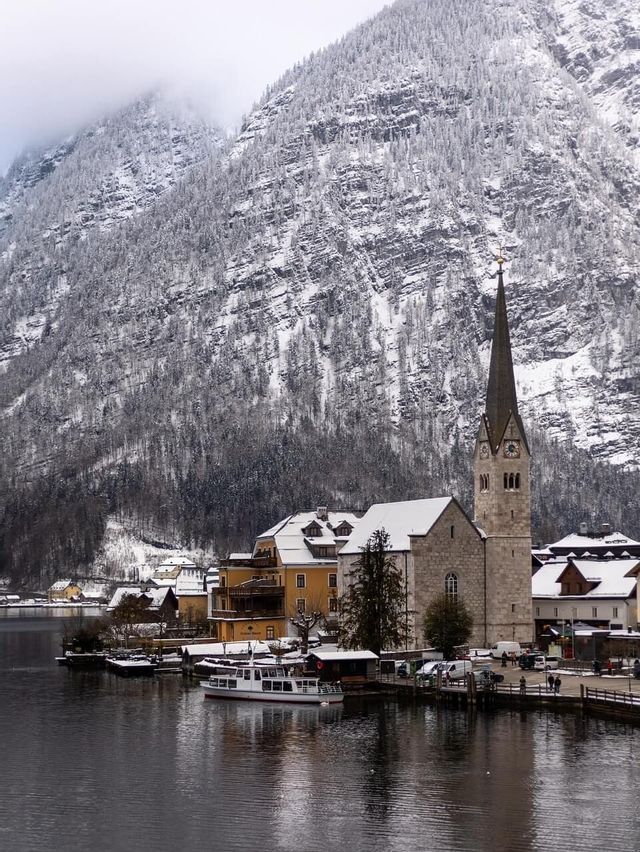 Am Fuße eines großen, schneebedeckten Berges liegt ein Dorf am Seeufer mit einer Kirche und einem hohen Kirchturm.