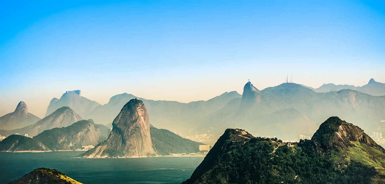 A wide view of mountains rising from the ocean, with a large statue visible on a distant peak under a clear blue sky.