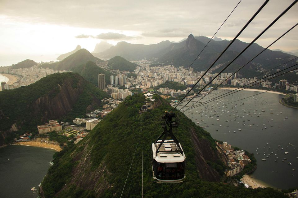 A cable car descends over a city surrounded by green mountains and a bay, with a monument visible on a distant peak.