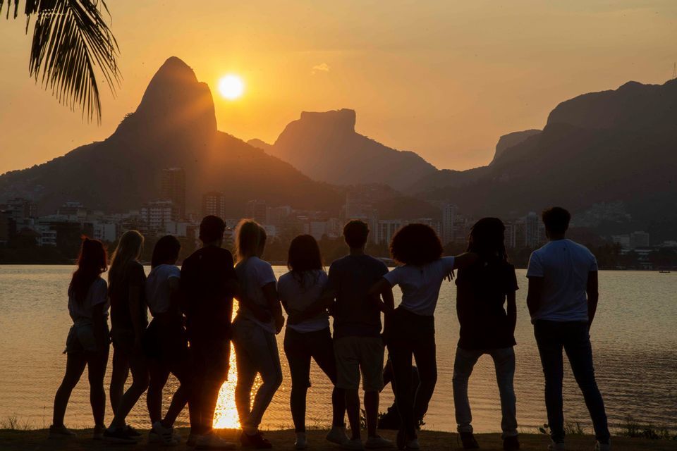 A WeRoad group trip silhouetted as they stand by a lake, watching the sunset over distant mountains and a city skyline.
