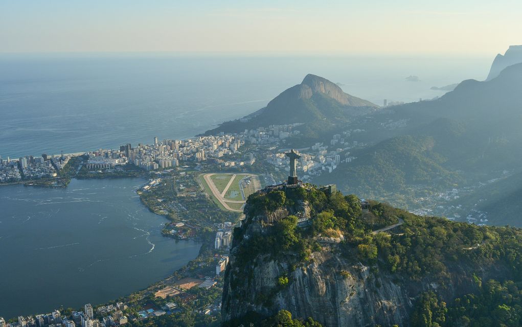 An aerial view of a large statue on a mountain peak, overlooking a coastal city with a large lagoon and the ocean in the background.