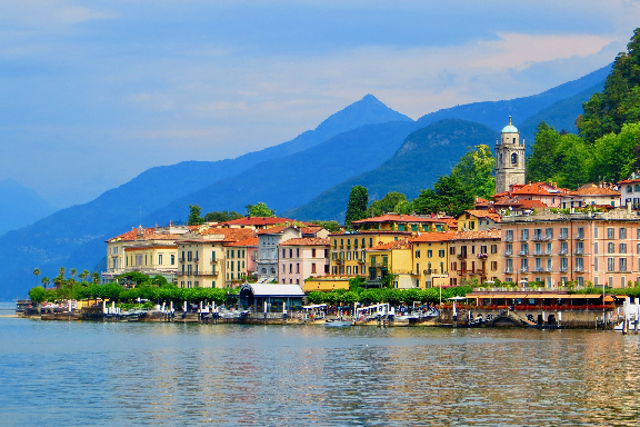 Una colorata cittadina in riva al lago con un campanile è incastonata ai piedi di grandi montagne alberate sotto un cielo azzurro.