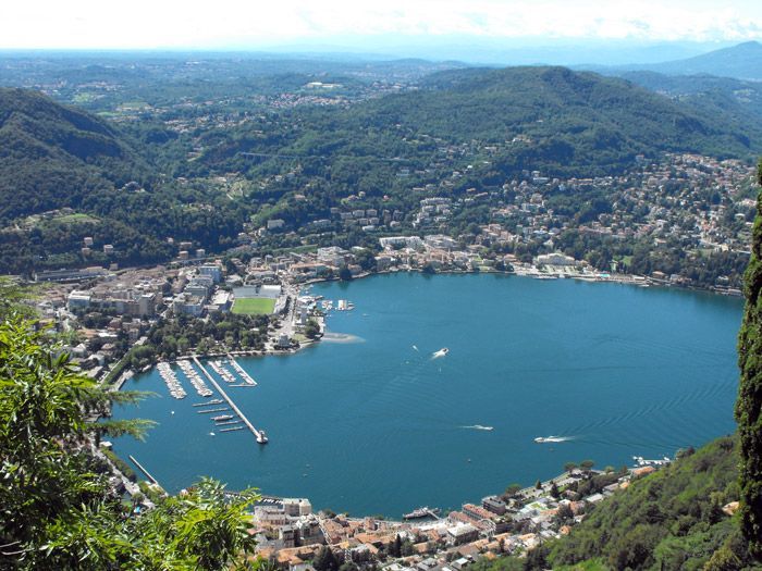 Una vista aerea di una città costiera con un porto turistico, incastonata tra un grande lago blu e rigogliose colline verdi.