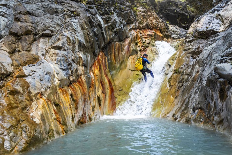 Une personne en combinaison de plongée et casque, avec un sac à dos jaune, descend en rappel une cascade dans un bassin d'eau situé dans un canyon rocheux.