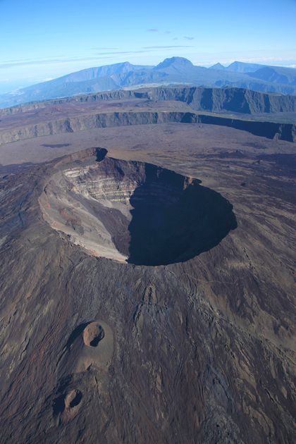 Vue aérienne d'un grand cratère volcanique dans un vaste paysage rocheux avec des montagnes lointaines sous un ciel bleu.