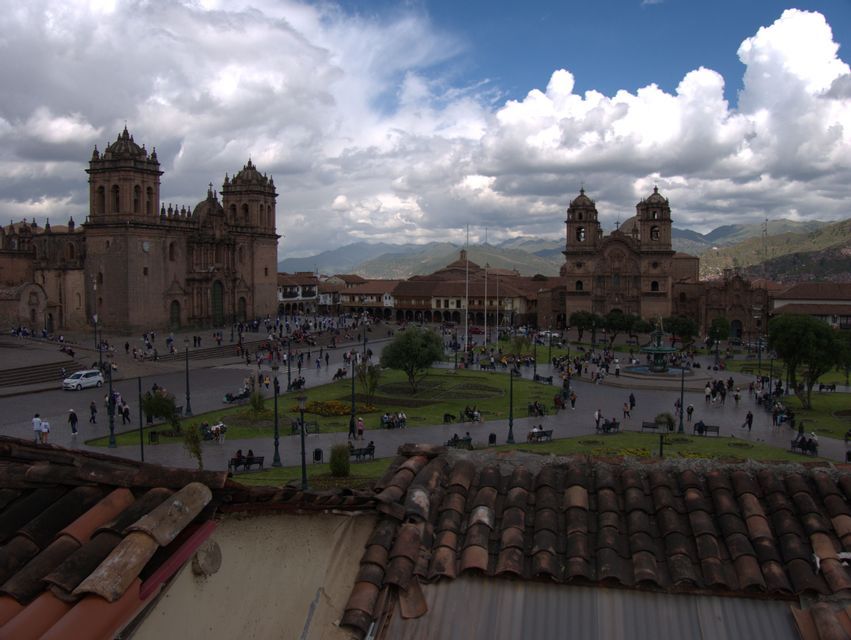 Ein erhöhter Blick auf einen belebten historischen Platz mit zwei großen Steinkathedralen, einem zentralen Park und Bergen im Hintergrund.