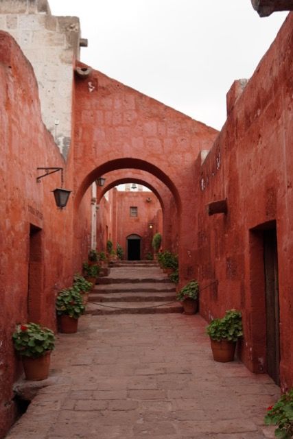 Eine schmale Steingasse mit terrakotta-roten Mauern und Bögen führt eine Treppe hinauf, gesäumt von Topfpflanzen.