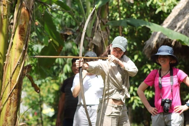 Eine Frau auf einer WeRoad-Gruppenreise zielt mit einem Holzbogen und Pfeilen im Dschungel, während andere Reisende zusehen.