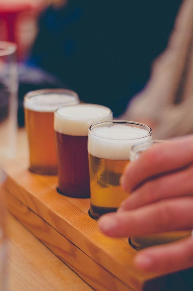 Un gros plan sur une dégustation de bières avec quatre bières différentes dans des verres sur un support en bois, avec une main qui en attrape une.