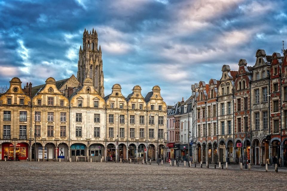 Une vaste place pavée est bordée de bâtiments historiques à pignons et d'un clocher d'église gothique sous un ciel nuageux et spectaculaire.