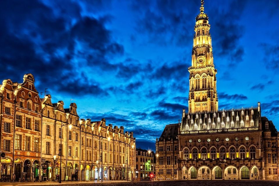 Un beffroi orné et des bâtiments historiques sont illuminés sur une place pavée au crépuscule sous un ciel d'un bleu profond.