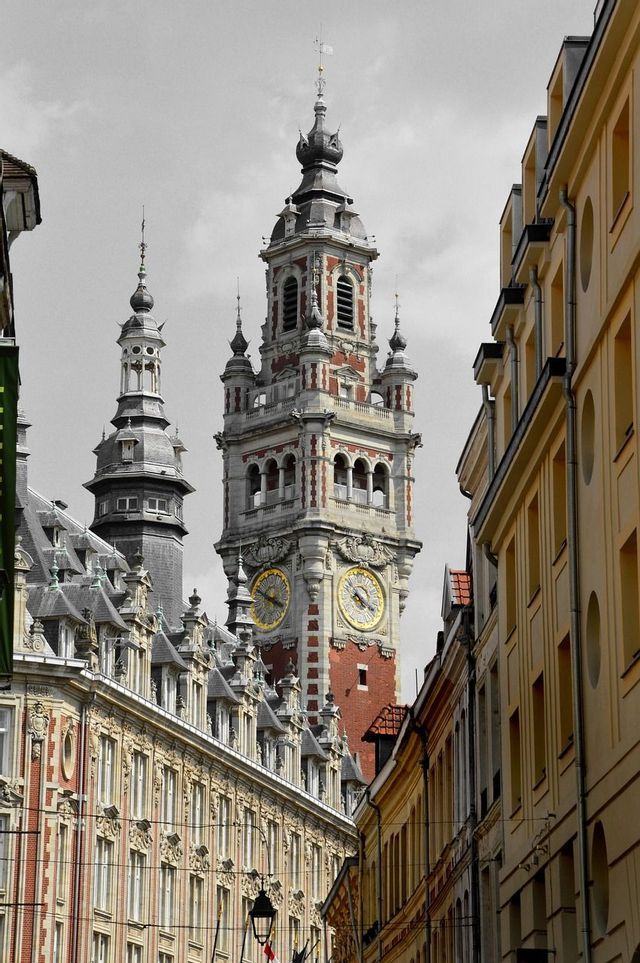 Une tour horloge ornée en briques rouges avec des cadrans dorés s'élève au-dessus d'une rue étroite bordée de bâtiments historiques, sur fond de ciel en noir et blanc.