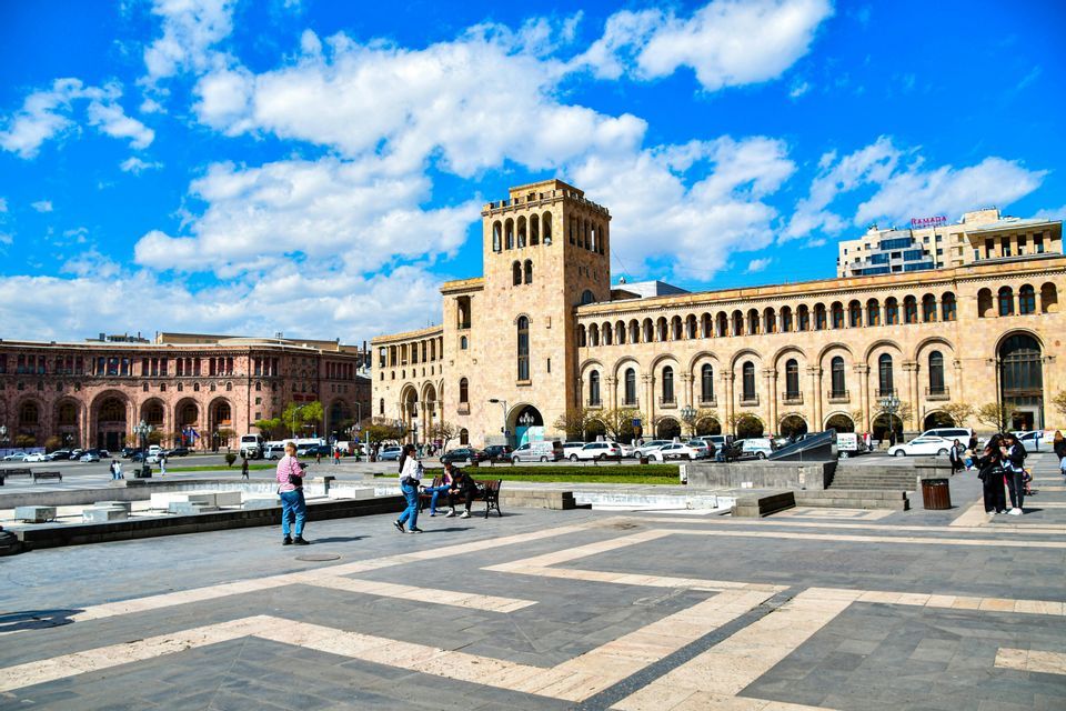 Un'ampia piazza in pietra decorata di fronte a imponenti edifici governativi ad arco, sotto un cielo azzurro brillante con soffici nuvole bianche.