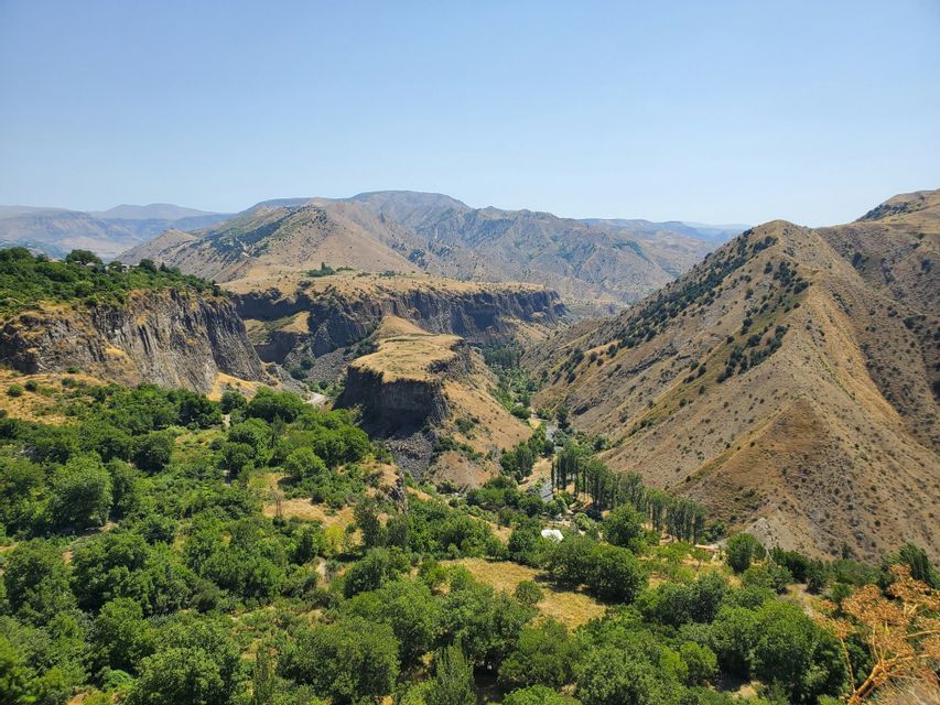 Una vista panoramica di un canyon profondo con un fondo valle lussureggiante e verde, circondato da montagne aride sotto un cielo sereno.
