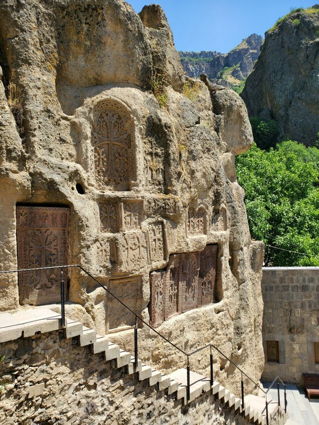 Intagli ornamentali a croce su una parete rocciosa accanto a una scala in pietra, con un canyon verde lussureggiante sullo sfondo.
